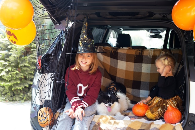 Photo two girls and dog celebrating halloween. safe halloween in quarantine. two sisters siblings sitting in car trunk with pet and having fun celebrating halloween. trick or treat. stay home