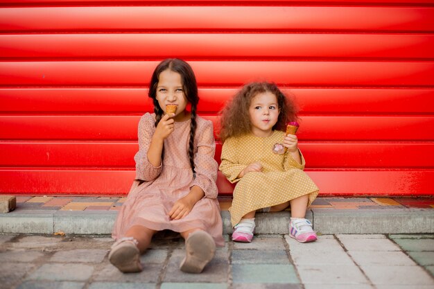Две девушки едят мороженое у красного забора. Одна брюнетка с двумя косичками, вторая - легкий кудрявый локон.