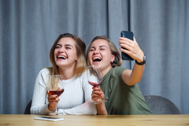 Two girls of European appearance sit at a table with glasses of wine and take a photo on the phone