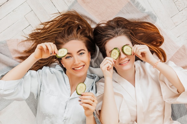 Two girls make homemade face and hair beauty masks. Cucumbers for the freshness of the skin around the eyes. Women take care of youthful skin. Girlfriends laugh at home lying on the floor on pillows.