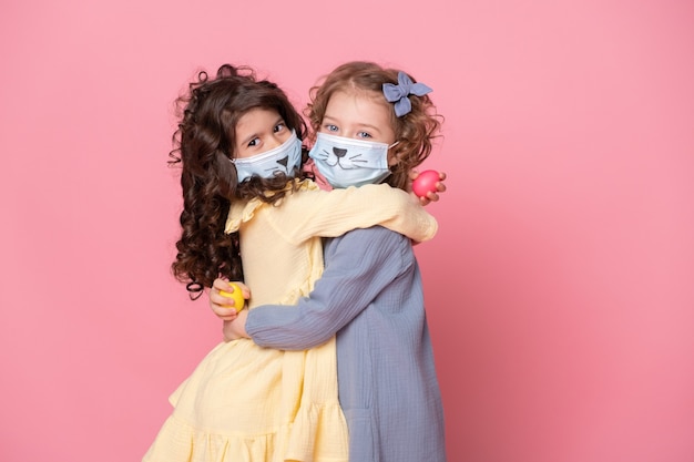 Two girls in protective mask with colored eggs on pink background. Covid easter concept.