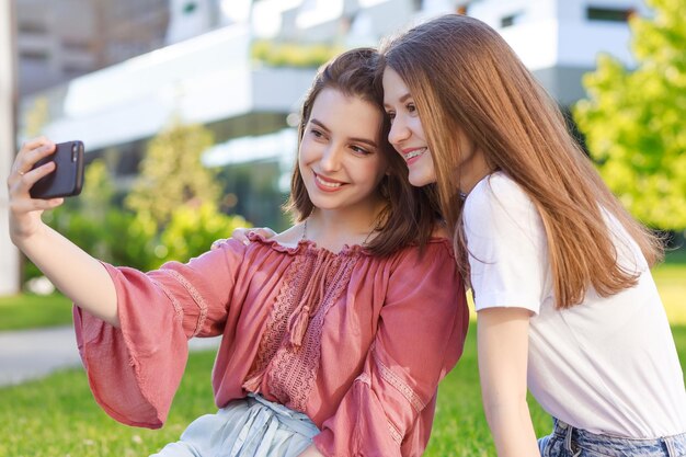 도시 공원에서 여름에 두 명의 여학생이 평상복을 입고 스마트폰으로 셀카를 찍습니다.
