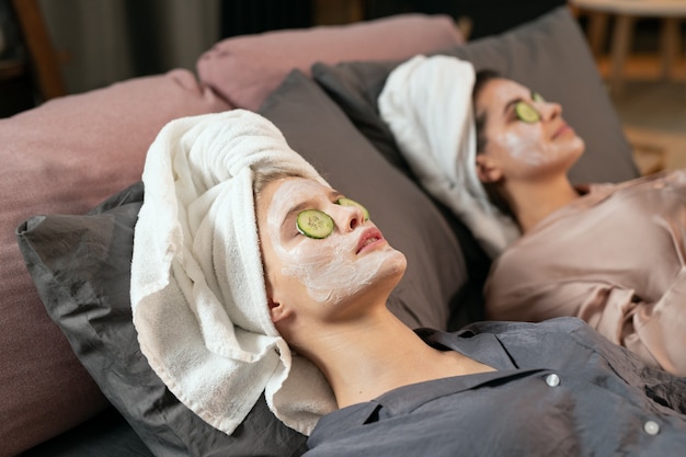 Две девушки в шелковой пижаме, полотенца на головах, глиняные маски на лицах и дольки огурца с закрытыми глазами, лежа на кровати и наслаждаясь процедурой красоты