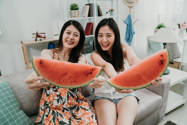 two girls sisters holding slices of watermelon in hands face camera showing smiling. Bright colours summer style healthy diet. women sitting on couch sofa laughing sharing fresh fruit in cozy home.