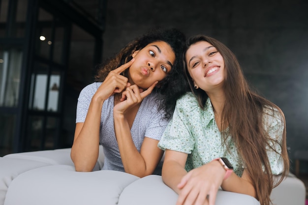 Photo two girls sit on the back of the sofa look at the cameras and smile