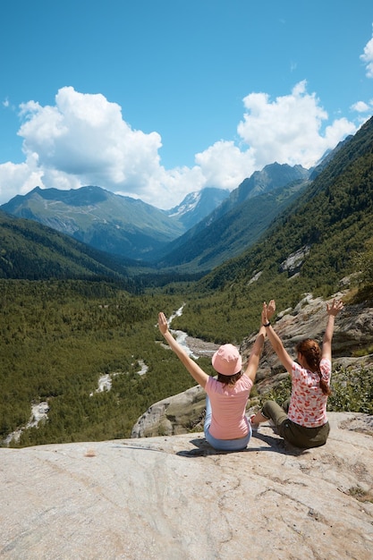 Two girls sits on the edge of a cliff and looks into the distance in the distance there is a river and a gorge a good mood clouds and a mountain landscape