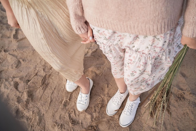 スニーカーと風通しの良いドレスを着た2人の女の子が、恋をしている女の子の脚を上から見た手を握っています...