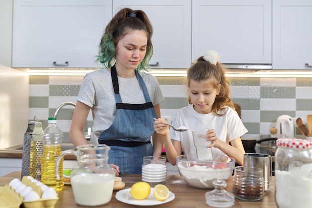 Two girls, teenager and younger sister, preparing cookies together in kitchen. Children mix flour in bowl, add ingredients. Family, friendship, fun, healthy homemade food