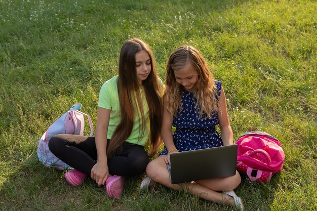 Two girls with laptop and briefcases doing homework or having fun while sitting on the grass outside