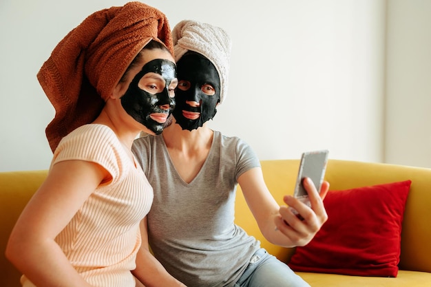 Two girls with a towel on their head and a black skin care mask they look in the mirror clean and pe...