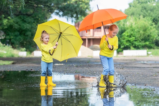 two girls in yellow clothes and with umbrellas merrily run through the puddles in rubber boots