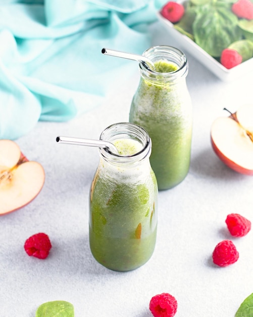 Two glass bottles of green smoothie with reusable drinking straws with spinach raspberry and apple on concrete table still life Healthy freshly blended smoothie for summer heat wave