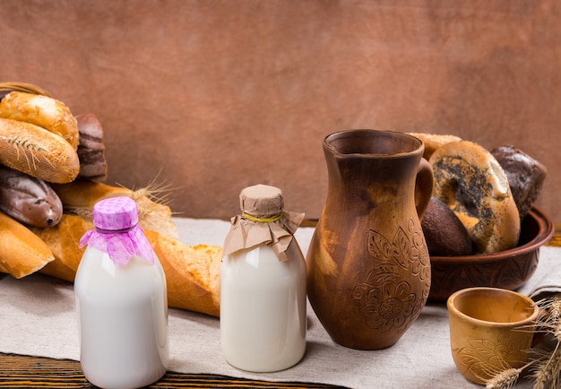 Two glass bottles of milk, tall wooden pitcher, little cup and bread loaves in background on table