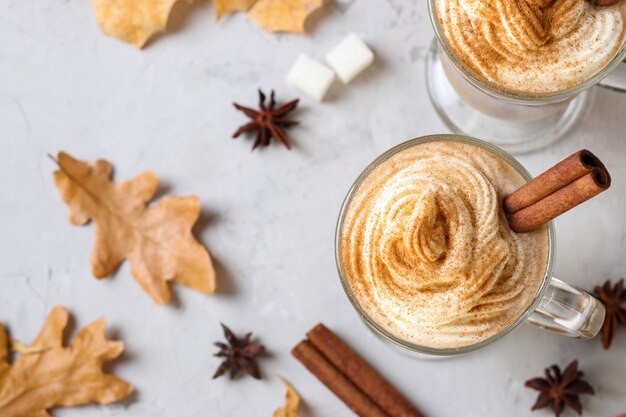 Two glass cups pumpkin latte with spices on grey background with autumn leaves. view from above