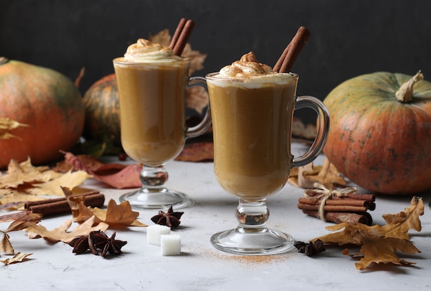 Two glass cups pumpkin latte with spices on grey surface with pumpkins and autumn leaves, close-up