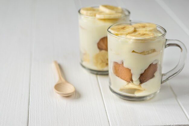 Two glass cups with banana pudding and wooden spoon on a white table. Milk and banana dessert.