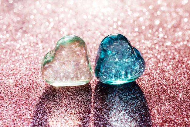 Two glass hearts, light green and blue on the blurry pink glitter background. Lights shines through transparent hearts.