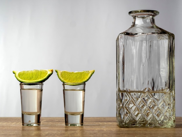 Due bicchieri di alcol con lime sulla parte superiore e una bottiglia di vetro su un tavolo di legno su sfondo bianco