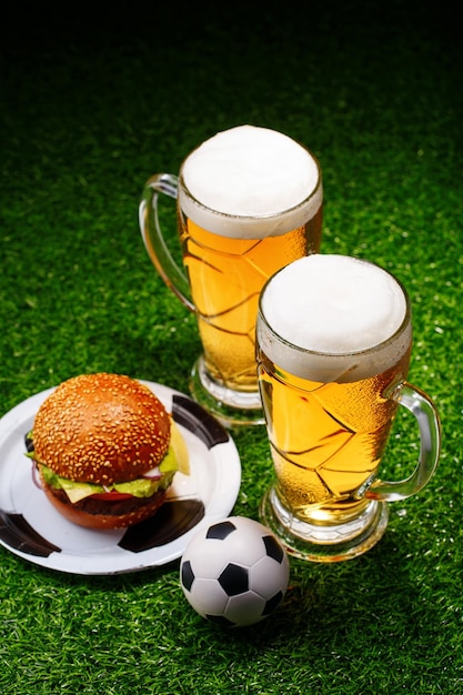 Two glasses of beer hamburger and soccer ball on green grass