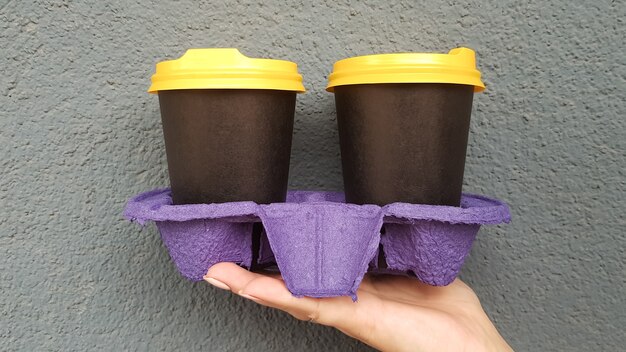 Two glasses of coffee in hand against the background of a blue wall. Takeaway coffee in disposable black cups with yellow lids. Morning coffee outside. Copy space.
