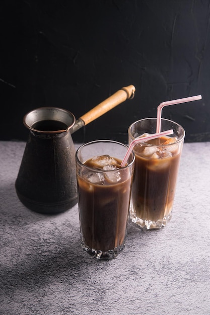 Two glasses of cold coffee with milk and ice with tubes on a dark background with a coffee maker