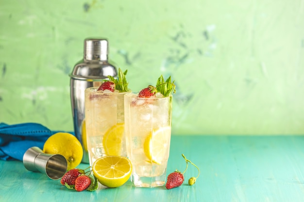 Two glasses of cold icy refreshing drink with lemon and strawberry served with bar tools on green wooden table.