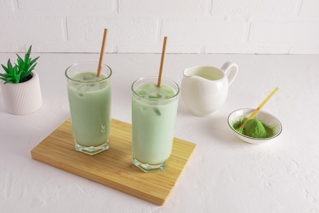 Two glasses of freshly made matcha tea and milk on a wooden board on a white table against a brick white wall an alternative to morning coffee