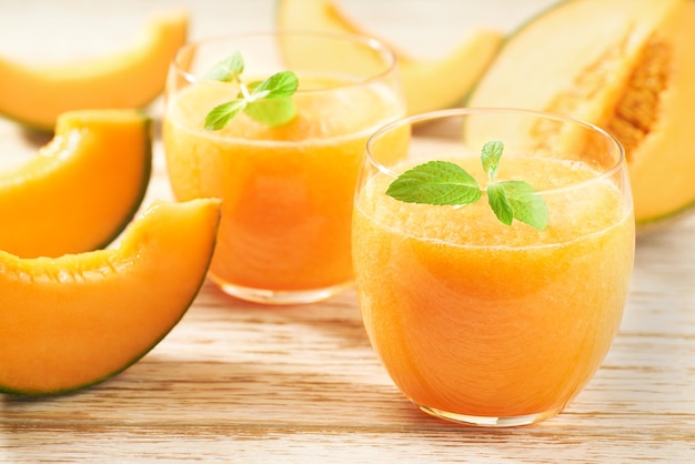 Two glasses of melon smoothie with fresh cantaloupe melon and mint on a wooden table.