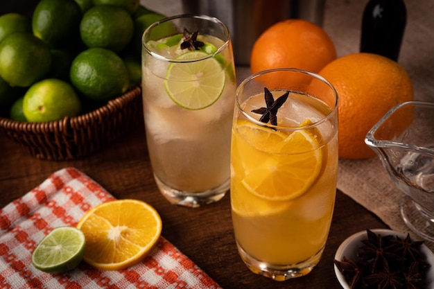 Two glasses of orange and lime cocktails sit on a table with a basket of oranges behind them.