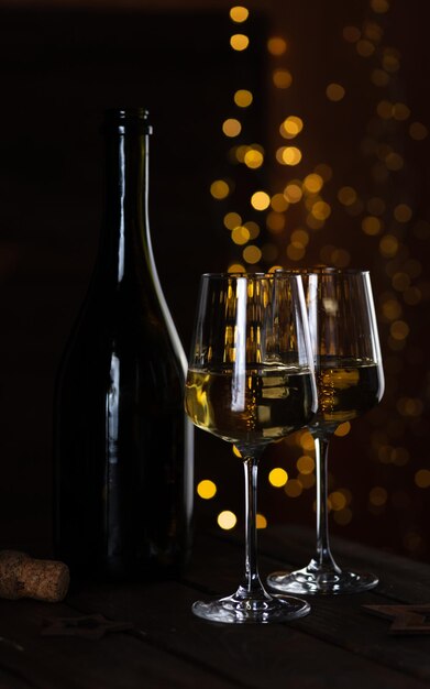 Two glasses of white wine and a bottle on a wooden table and lights garlands behind