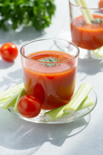 Два стакана со свежим томатным соком, сельдереем, петрушкой и спелыми помидорами на светлом фоне
