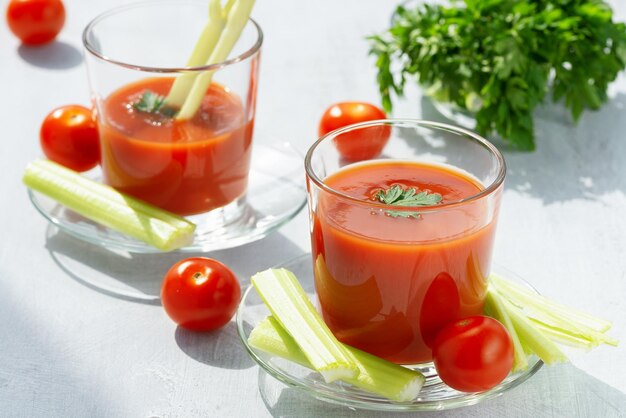 Two glasses with fresh tomato juice celery parsley and ripe\
tomatoes on light wood background