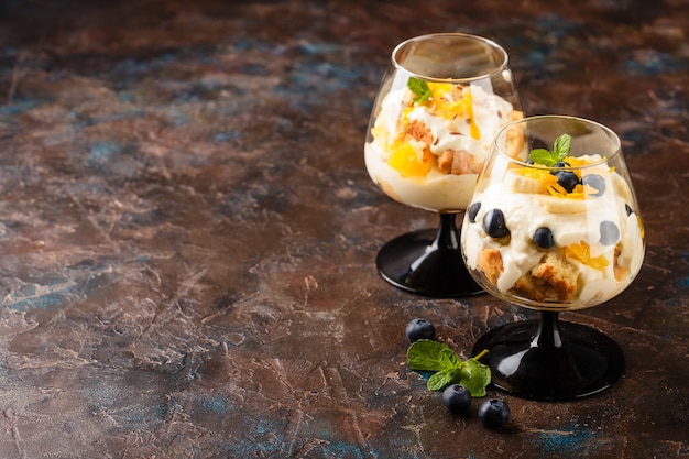 Two glasses with Healthy homemade layered dessert trifle with orange, blueberry, biscuit, yogurt and granola. Place for text