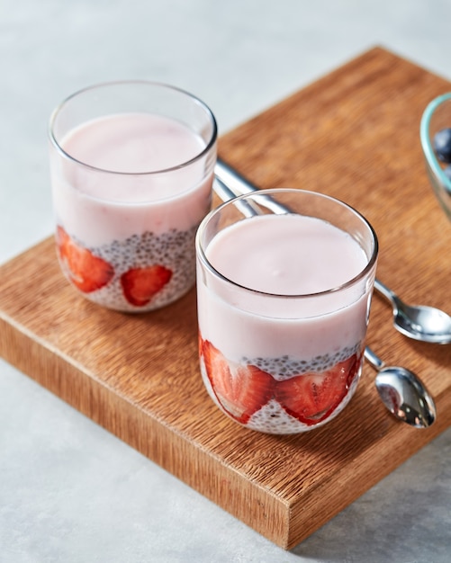 Two glasses with smoothie and pieces of srtawberries, chia seeds on a wooden board on a gray background. Concept of healthy dieting food.