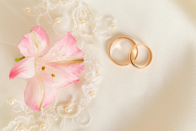 베이지색 새틴 배경 결혼식 배경 복사 공간에 두 개의 금 약혼 반지와 분홍색 아스트로메리아 꽃