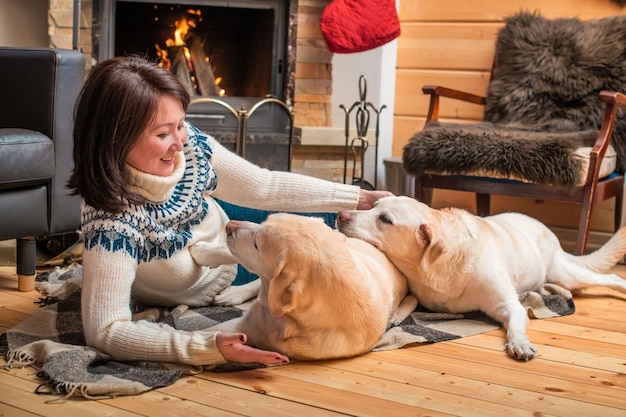 Two golden Labrador retriever dogs lie with a middle-aged Asian woman on a blanket in front of a country house fireplace.