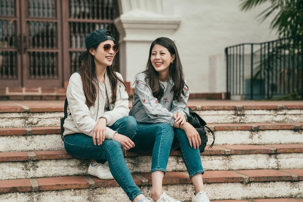 two good friends siiting on the stairs and laughing happily while traveling and hanging out together.