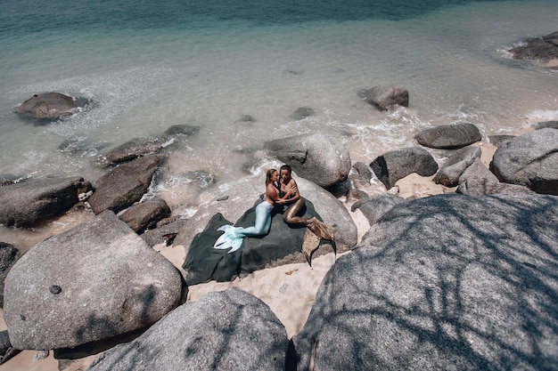 Two gorgeous lesbian mermaids with long tails are lying on the rocks and posing on the beach, stones and ocean