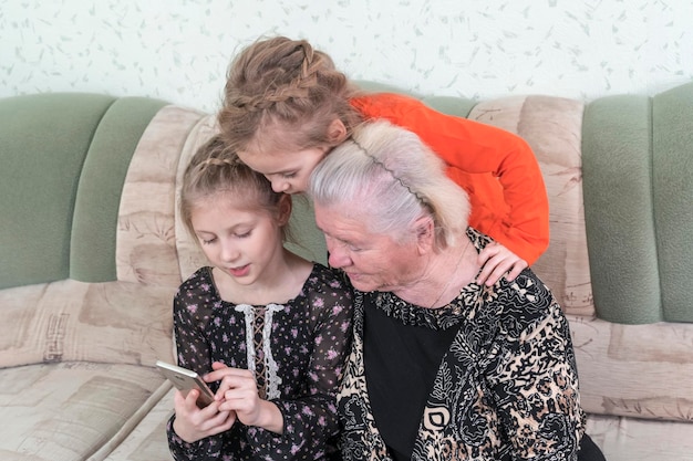 two granddaughters teach great-grandmother to use a smartphone while sitting on the couch