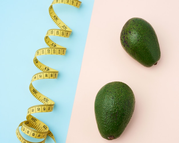Two green avocado fruits and a twisted measuring yellow tape on a beige-blue background