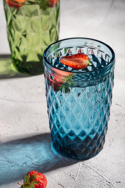 Two green and blue geometric glass cup with fresh water and strawberry fruits with colorful shadow light rays on stone concrete surface