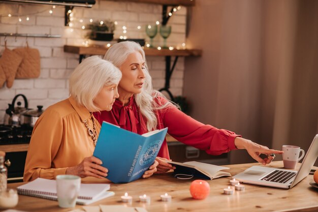 Two grey-haired pleasant cute ladies looking amused while studying together