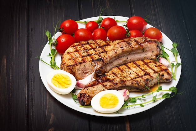 Two grilled pork steaks on a white plate with cherry tomatoes, sprouted chickpeas and boiled egg.