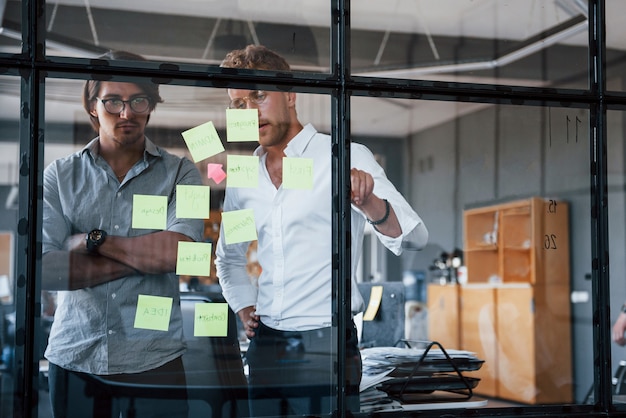 Two guys in formal clothes stands in the office behind the glass and puts stickers on it.