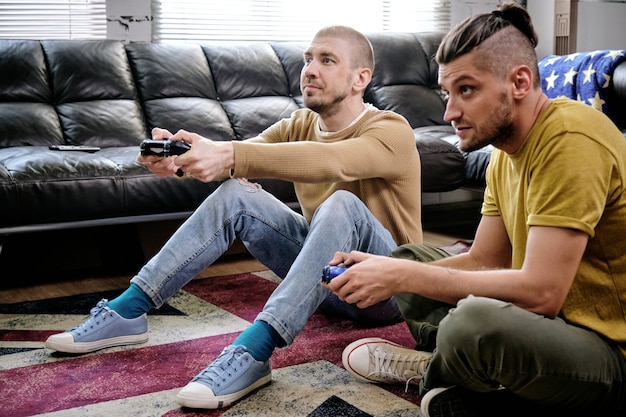 Two guys with joysticks playing video game on the floor of living-room