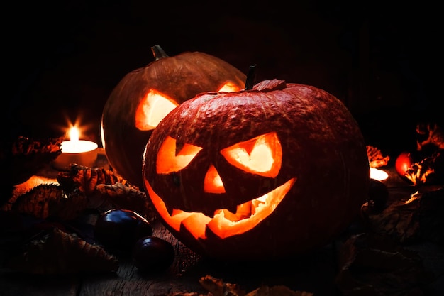 Two Halloween pumpkin JackoLantern on dark wooden background with fallen leaves and flames selective focus