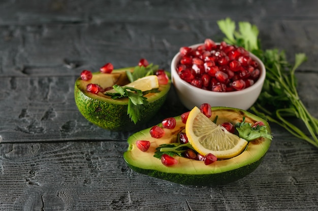 Two halves of avocado, ripe pomegranate, lemon and parsley leaves on a rustic dark wooden table.