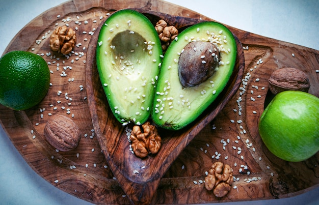 Two halves of avocado on the wooden cutting board with lime, apple and walnuts.