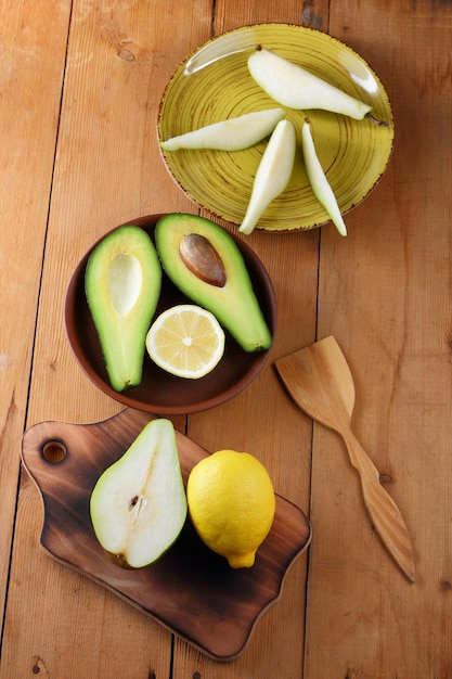 Two halves of a ripe avocado with seed on a plate Fresh green fruits with yellow lemon and wooden cutlery on wooden background Closeup View from above