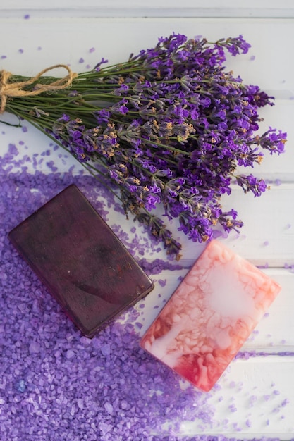 Two handmade soaps laying on white wooden table and violet sea salt near bouquet of fresh lavender flowers.